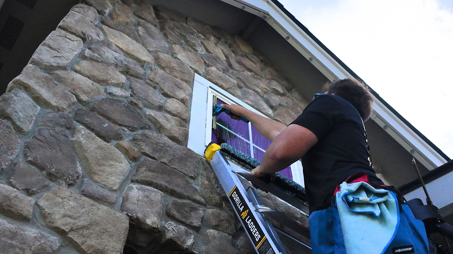 Technician on a Gorilla ladder reaching a high window on a stone-clad home